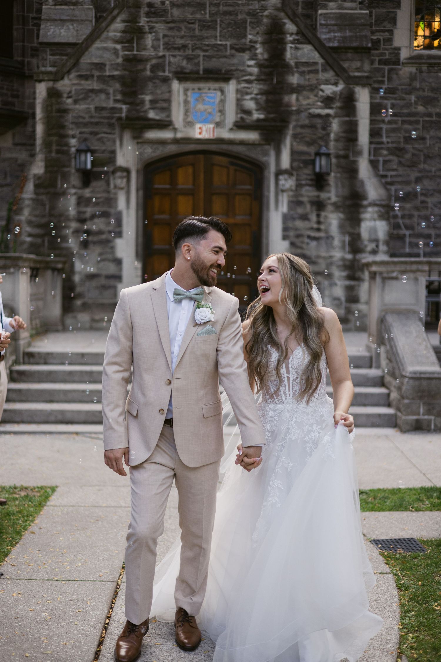 Bride and groom laugh together during romantic bubble exit outside historic stone wedding venue.