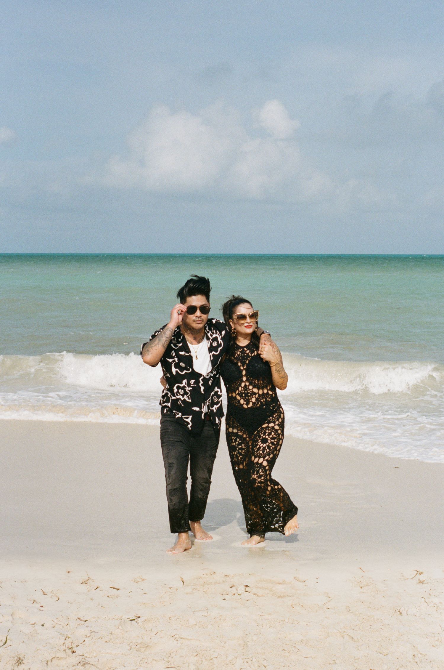 Stylish couple walking on a white sandy beach, wearing matching black outfits with sunglasses.