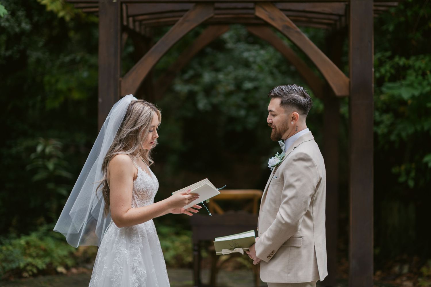Bride reading vows to groom in tan suit under a wooden arch in a lush garden ceremony.