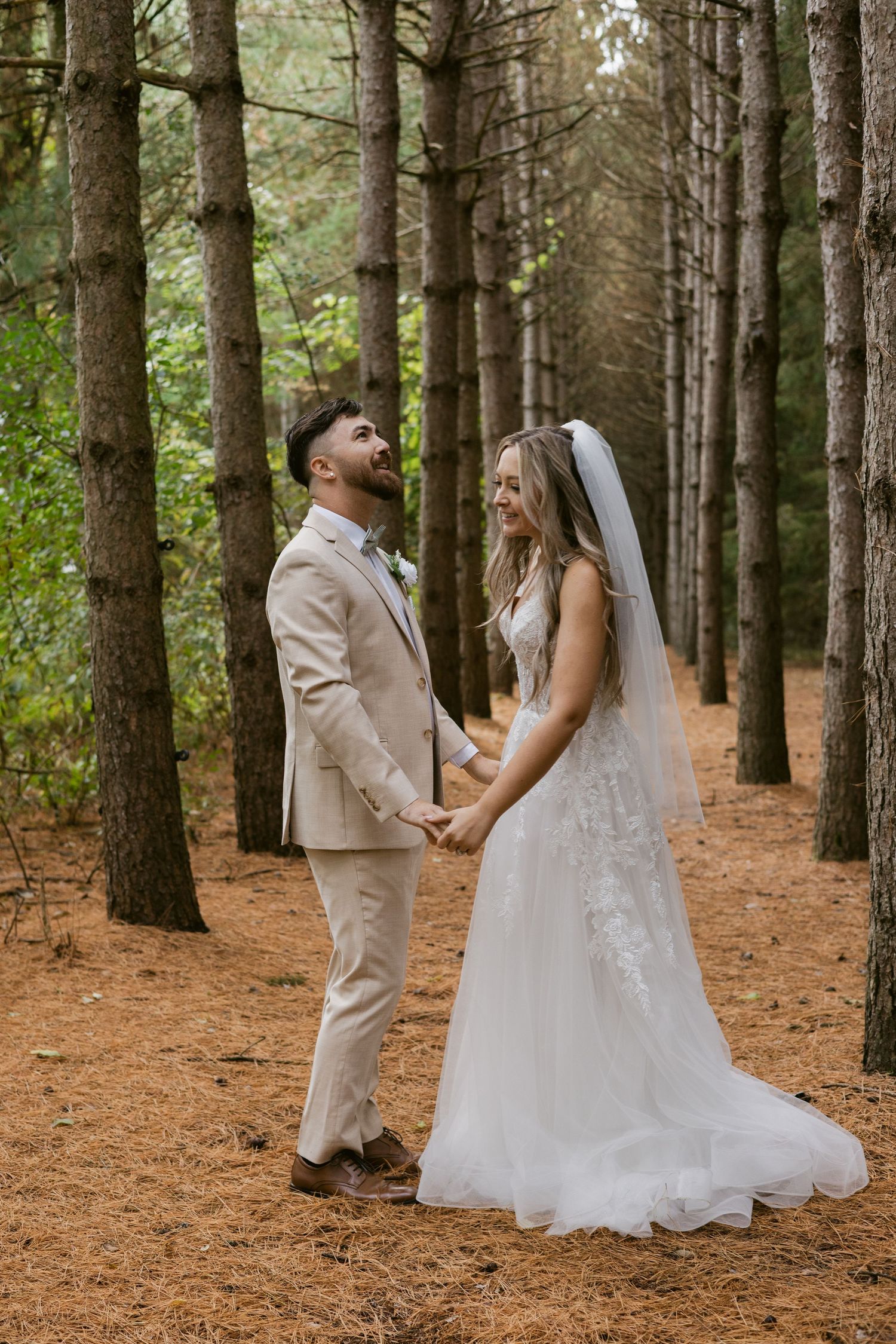 Bride and groom hold hands smiling in a pine forest, she in a lace gown with veil, he in a beige suit.