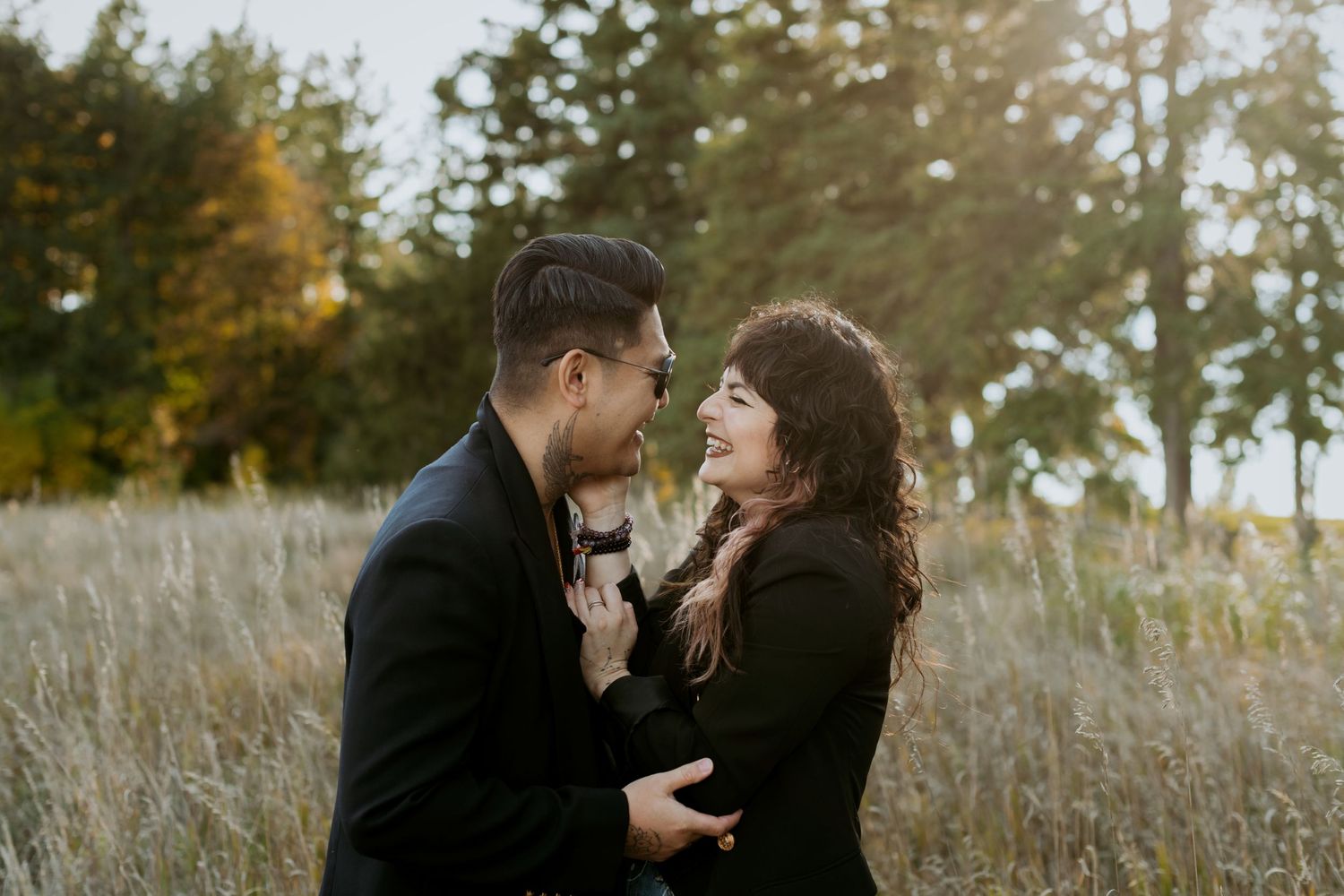 A romantic couple embracing face to face in a tall grass field with autumn trees behind them.