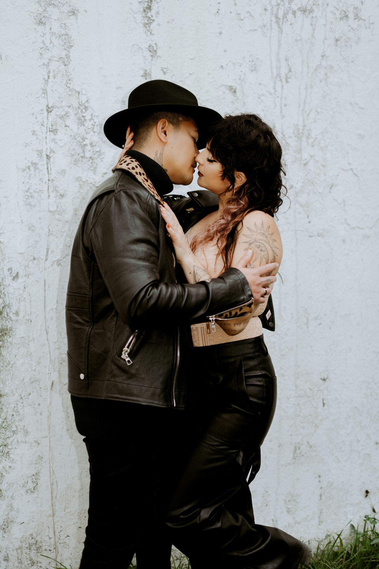 Couple in black leather jackets sharing a kiss against a rustic white wall.