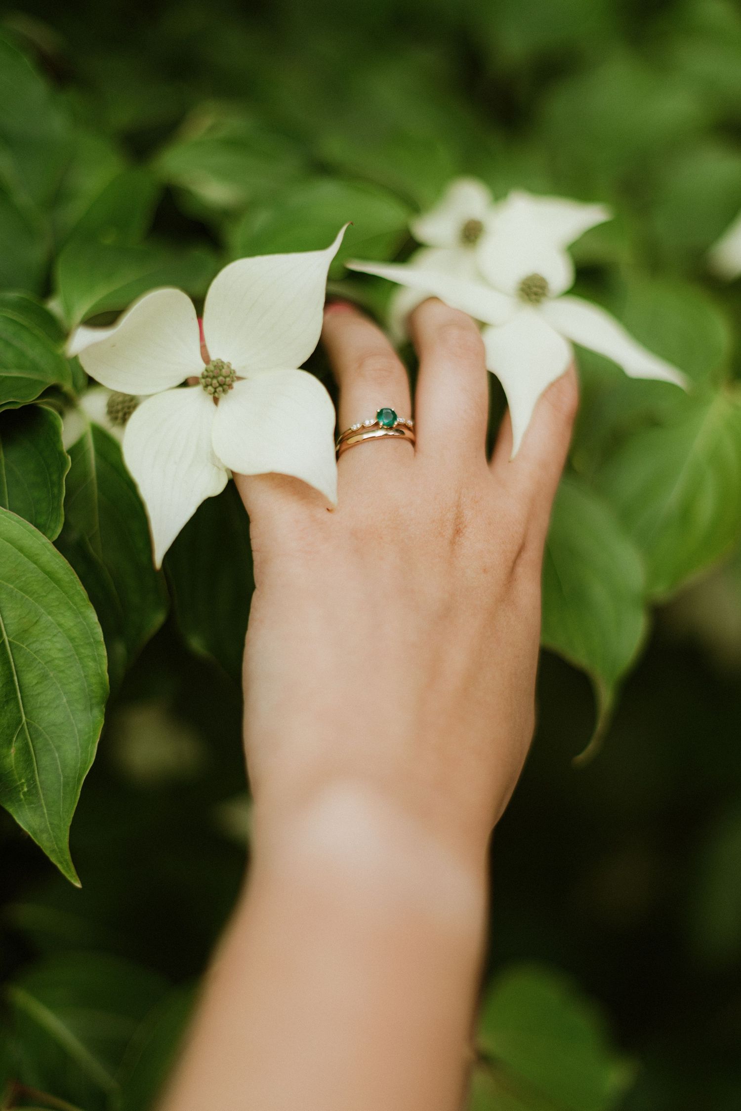 Close-up of bride's hand with emerald engagement ring and gold band touching white flowers.