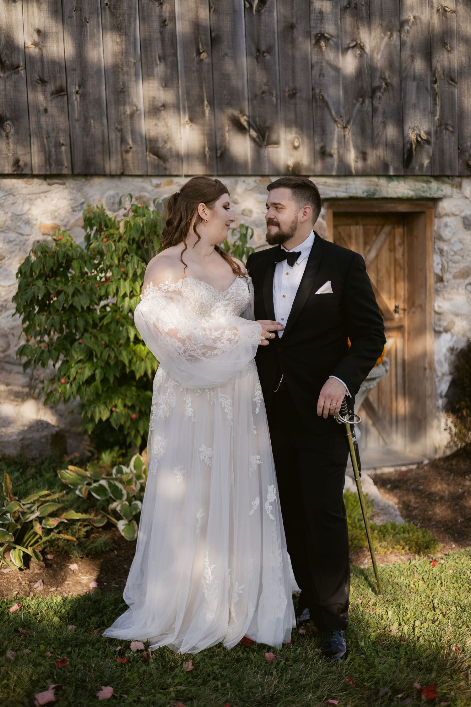 Plus-size bride in floral lace gown with groom in black tuxedo sharing romantic moment outside stone barn venue.