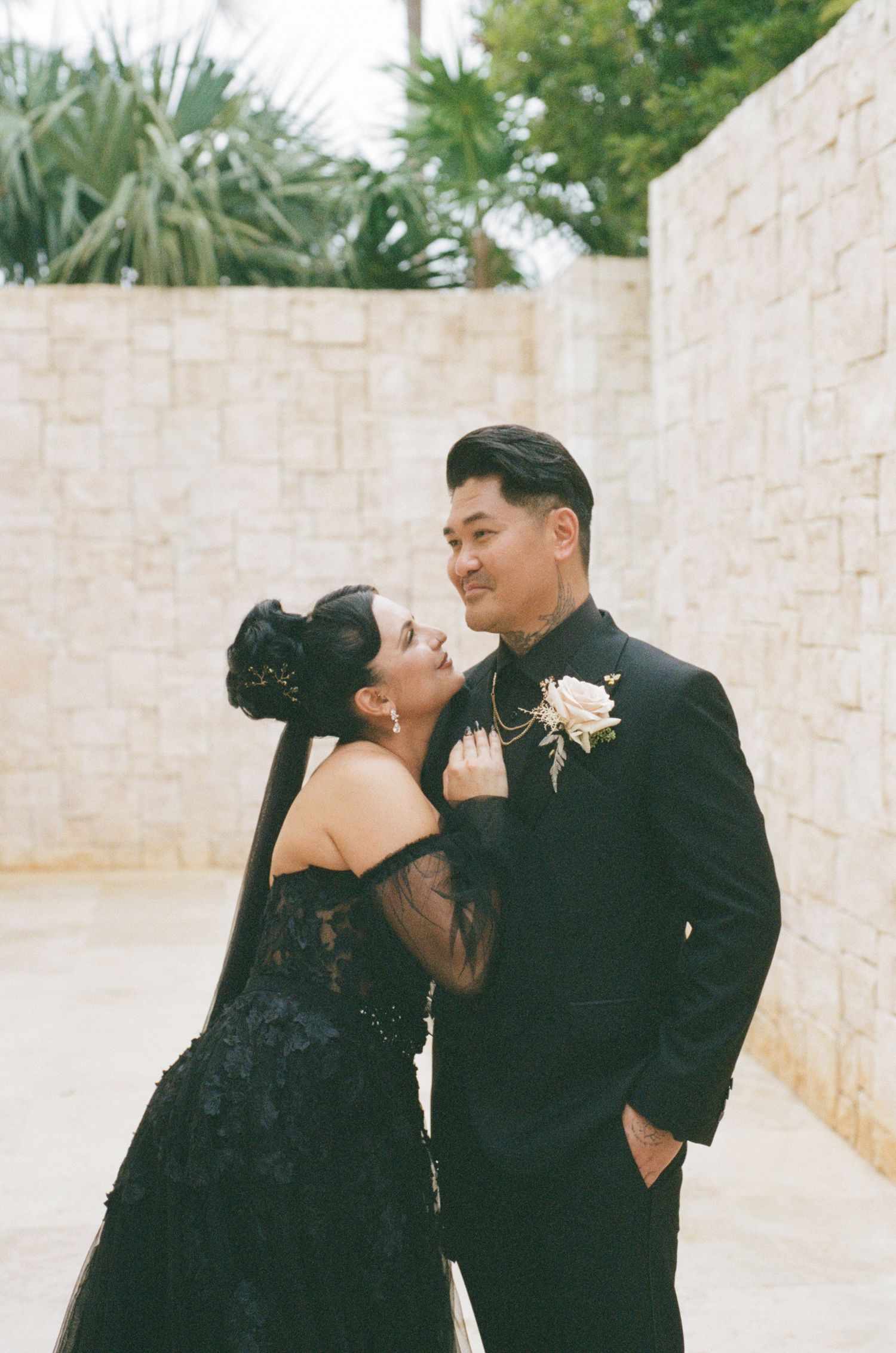 Bride and groom in black wedding attire sharing romantic gaze in stone courtyard setting.