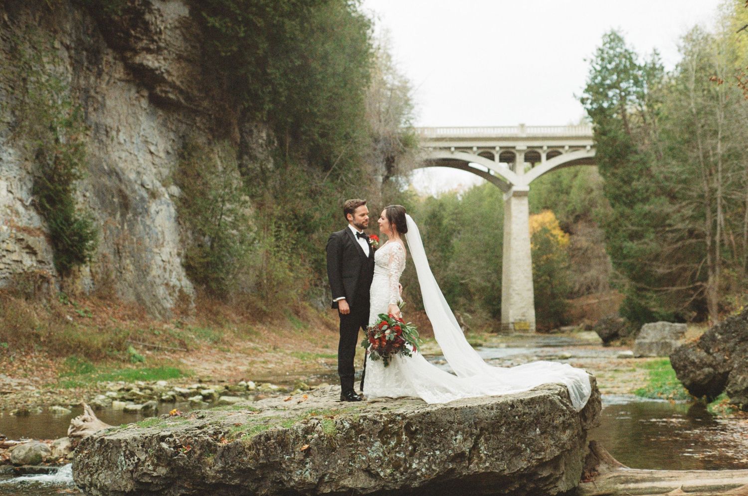 Bride and groom kissing on a rock by a river with a stone arch bridge in the background.