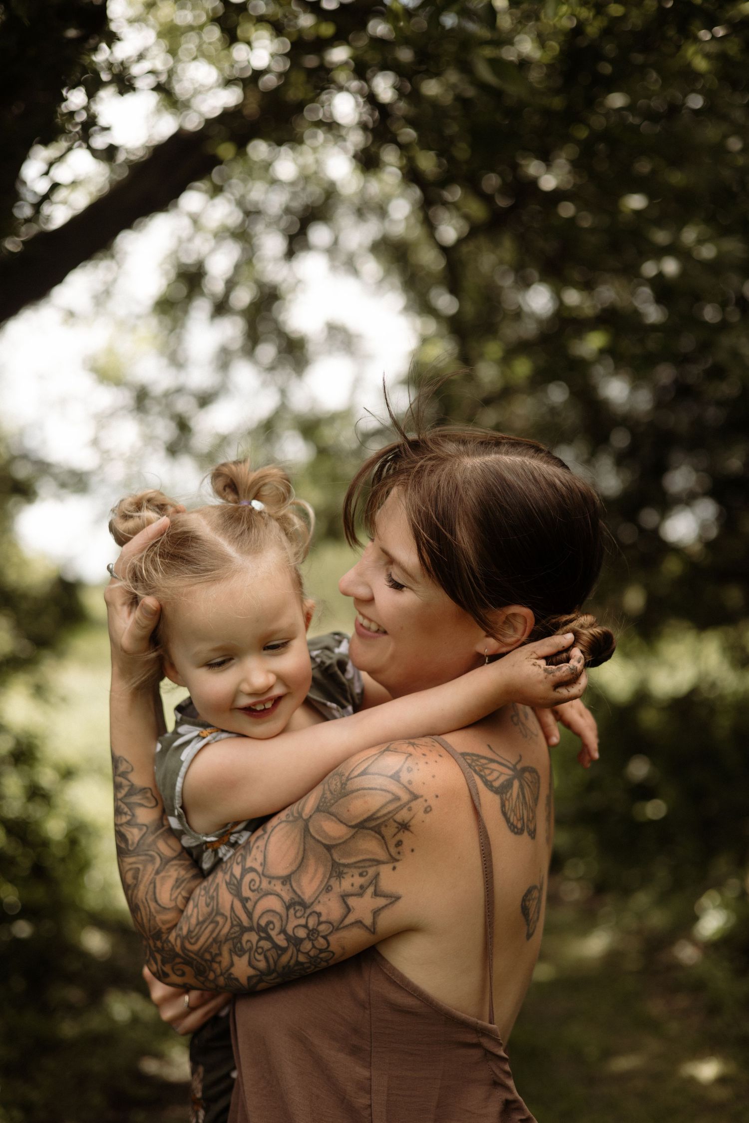 A tattooed mother smiles warmly while holding her happy toddler in a green park.