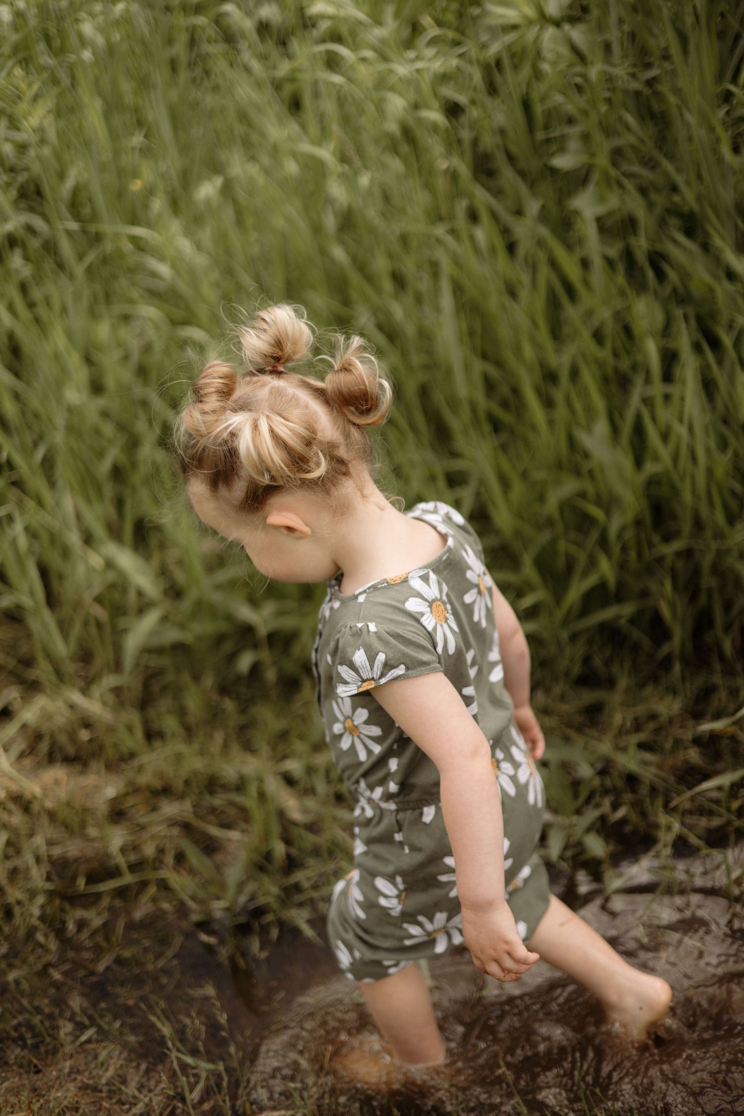 A cute toddler girl in a green daisy romper splashes barefoot in a muddy puddle near tall grass.