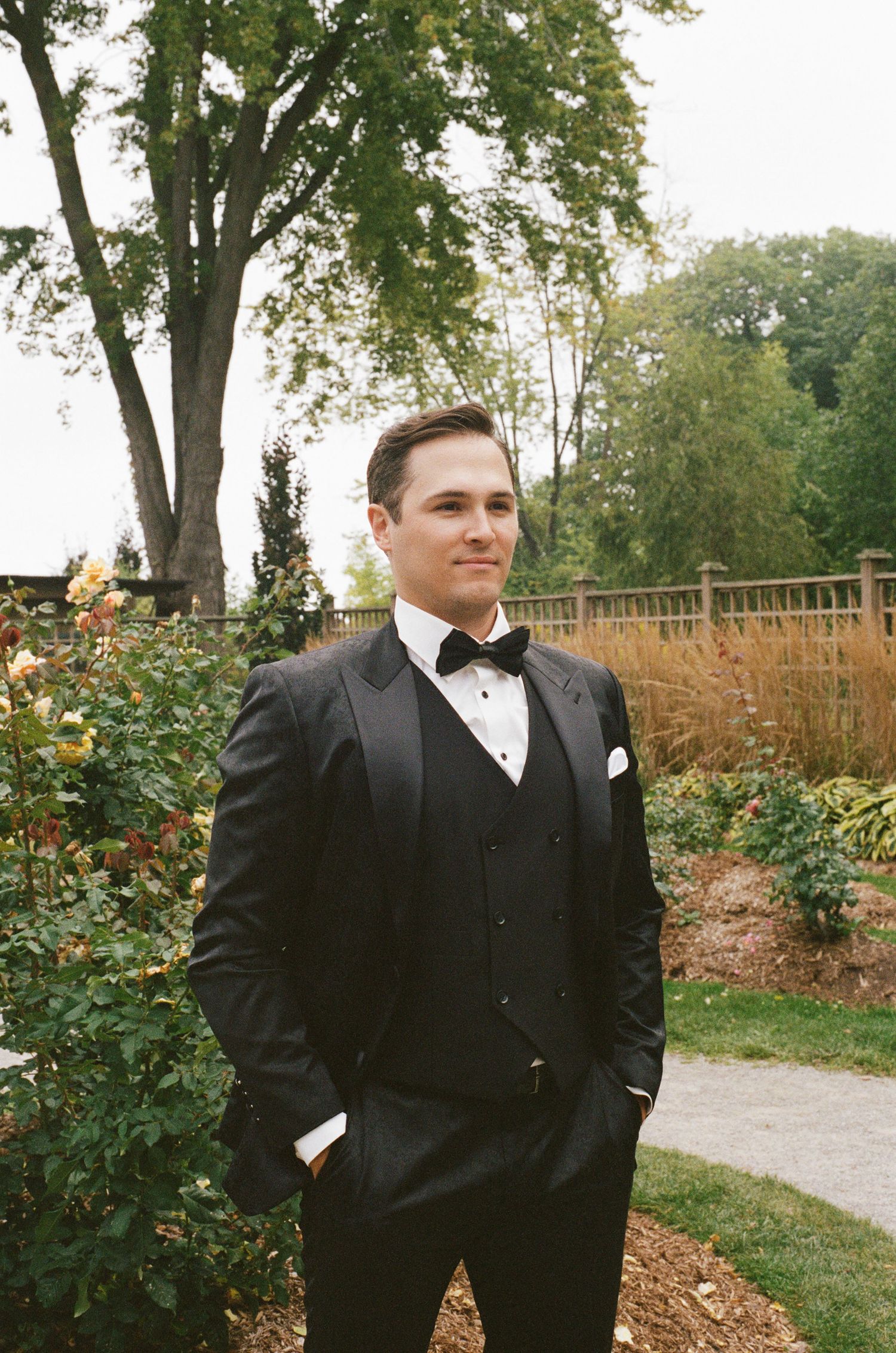 Dapper groom in black tuxedo with bow tie standing in a colorful garden setting.