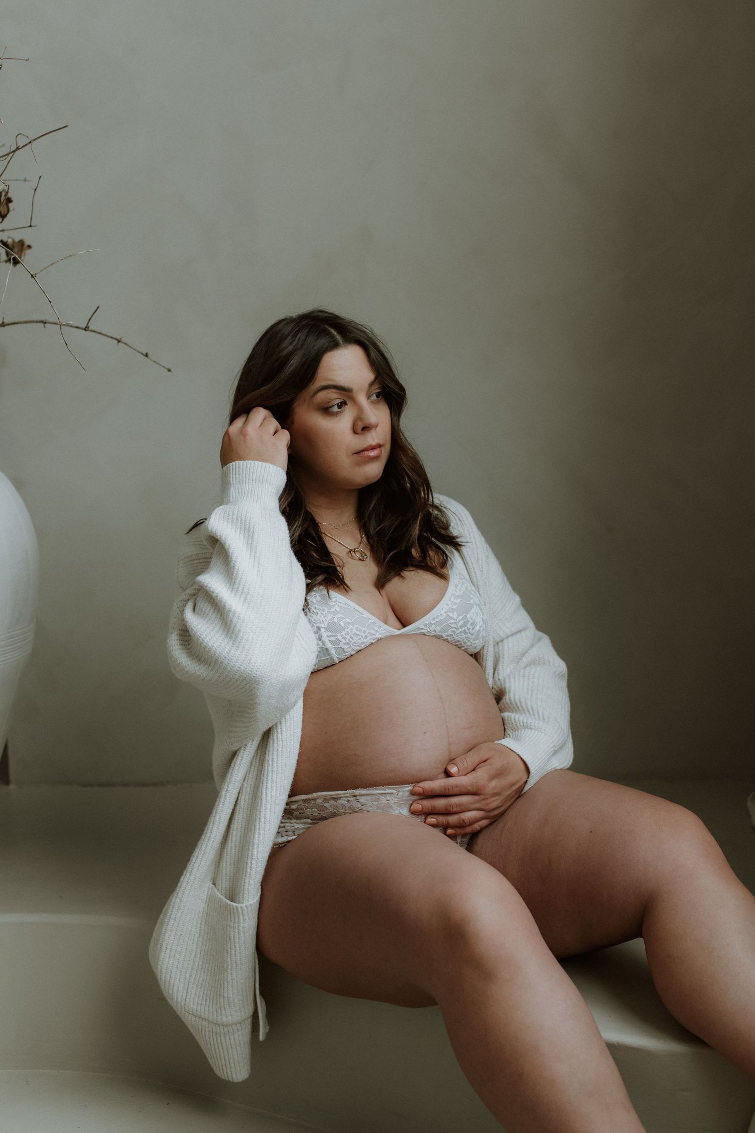 Pregnant woman in white lace lingerie and oversized cardigan sits gracefully in a neutral studio setting.