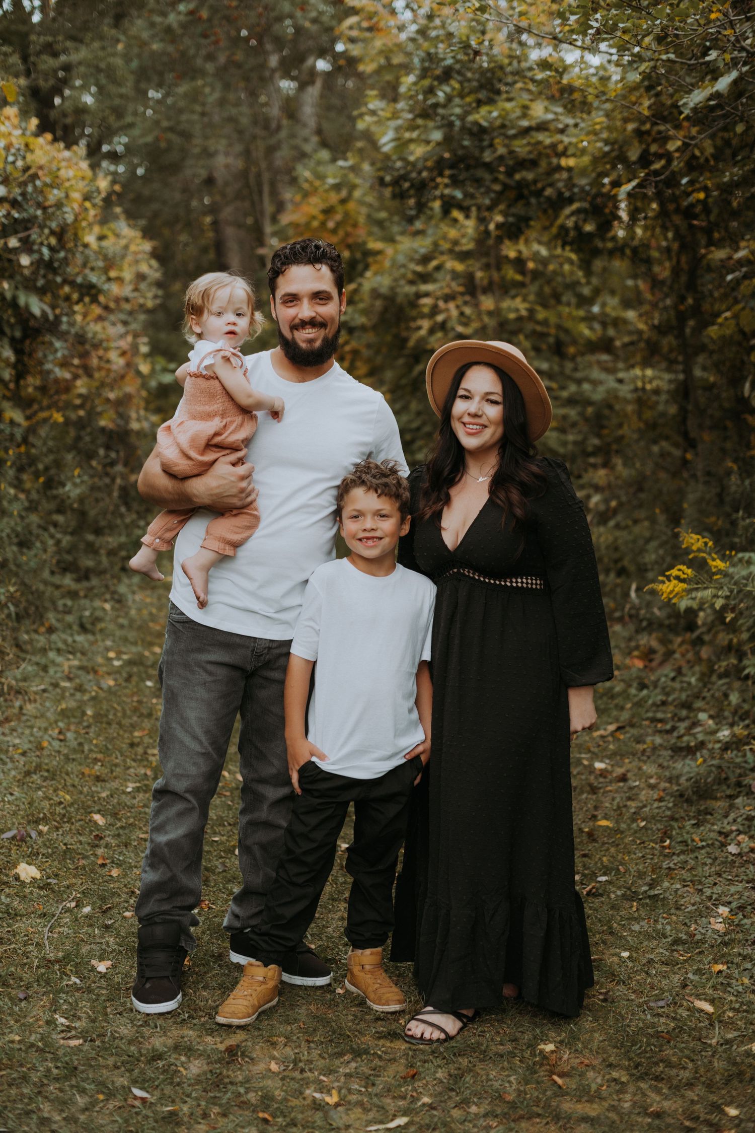 Family of four smiling together in autumn forest, father holding toddler in terracotta outfit.