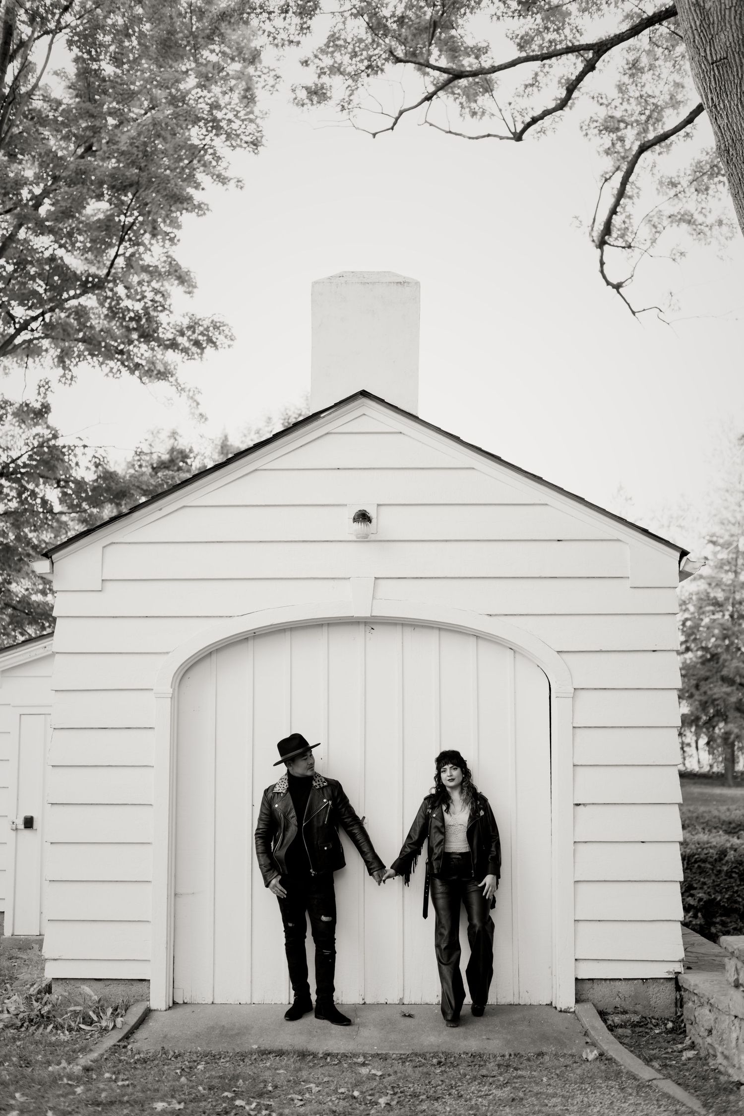 Black and white photo of a couple in leather jackets holding hands in front of a white barn door.