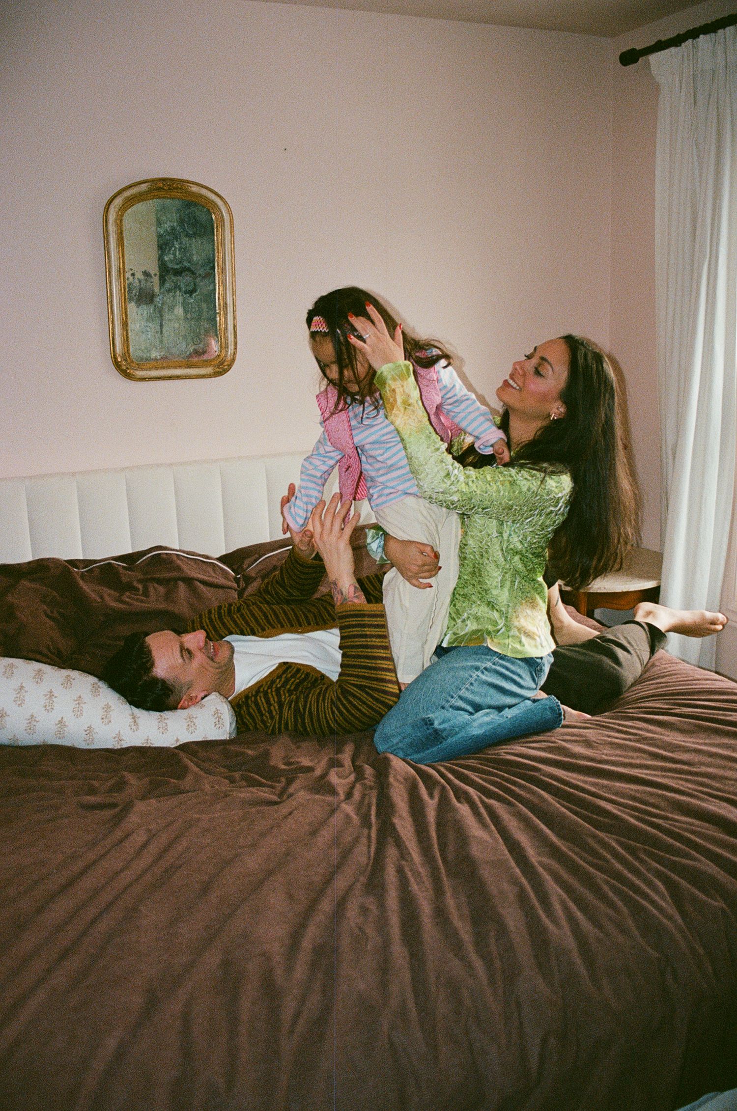 A joyful family plays together on a brown bed, with a woman lifting a child while a man lies smiling.