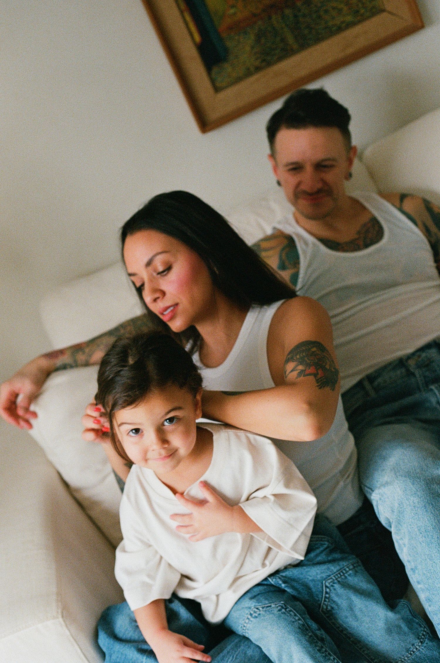 Smiling tattooed mother embracing young child on floor while father relaxes on white sofa.