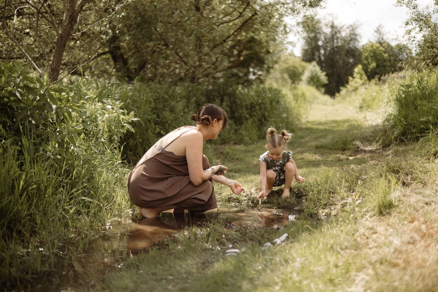 Mother and toddler sitting together by a small stream surrounded by lush green vegetation on a sunny day.