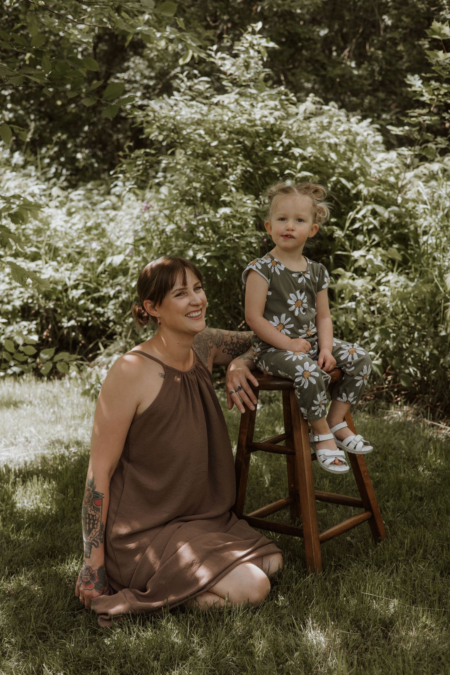 Tattooed mother in brown dress sitting on grass beside toddler perched on wooden stool outdoors.
