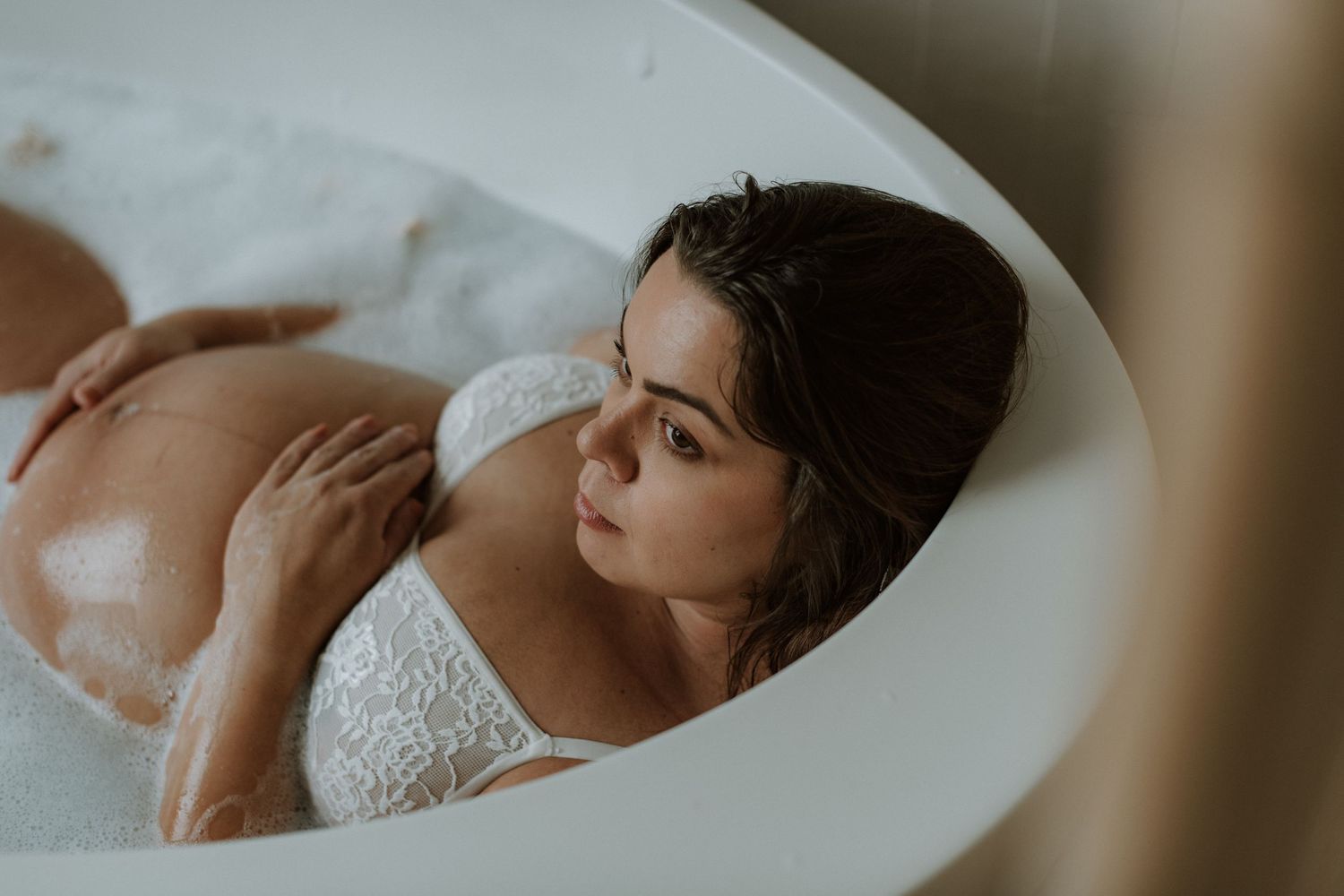 Pregnant woman in white lace bra relaxing in a milk bath, gently touching her belly.