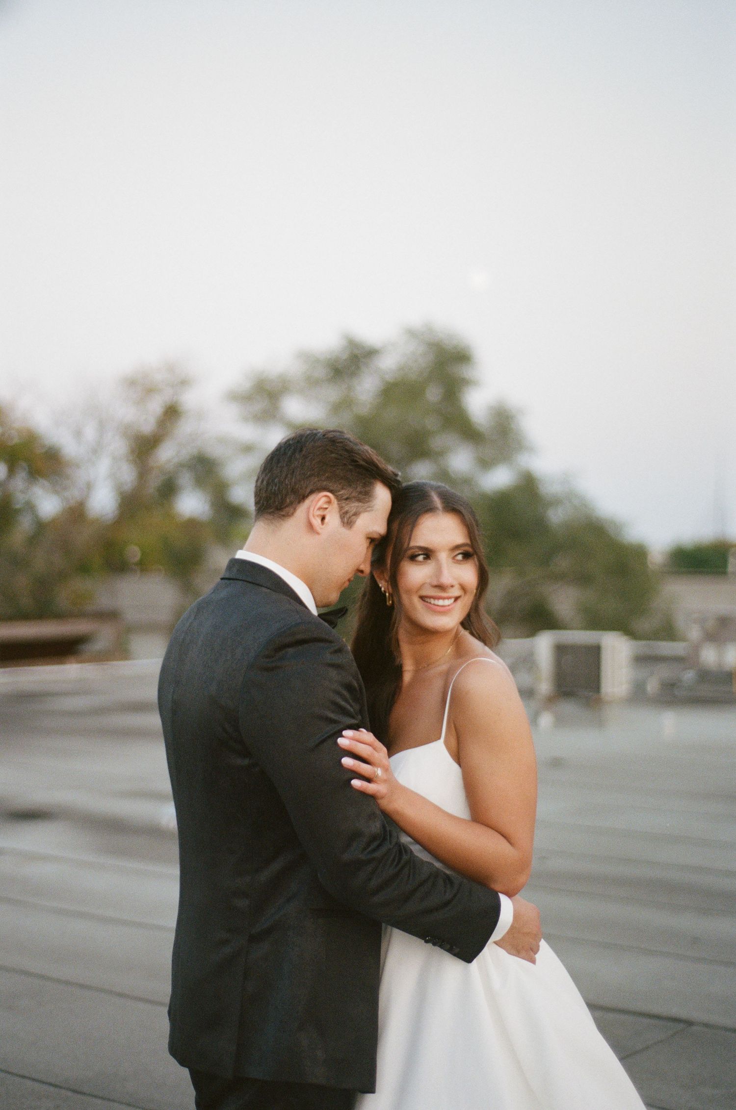 Radiant bride smiles over shoulder while groom embraces her tenderly on rooftop wedding location.