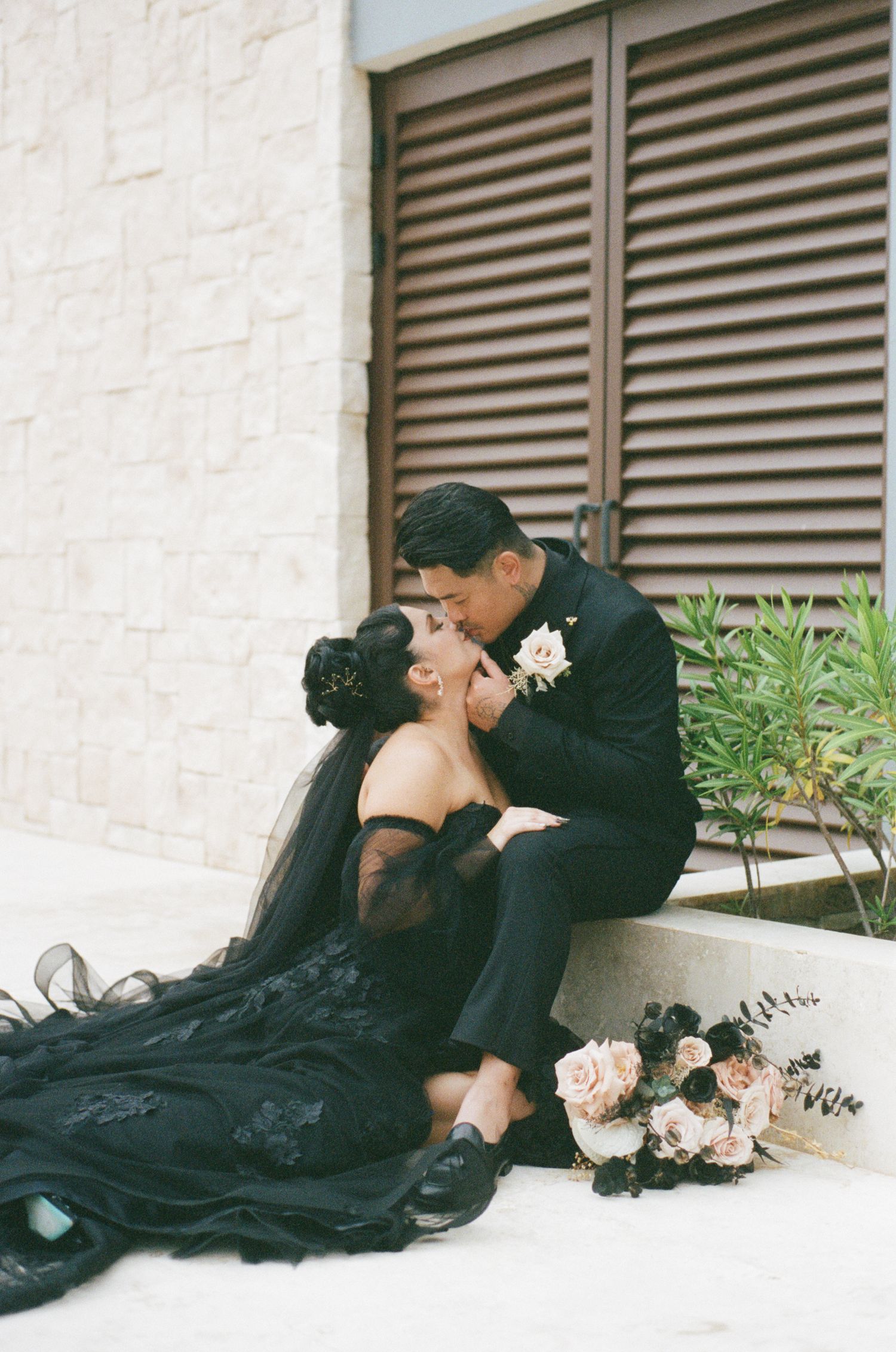 Bride and groom share romantic kiss while seated on steps, bride in black lace gown with bouquet.