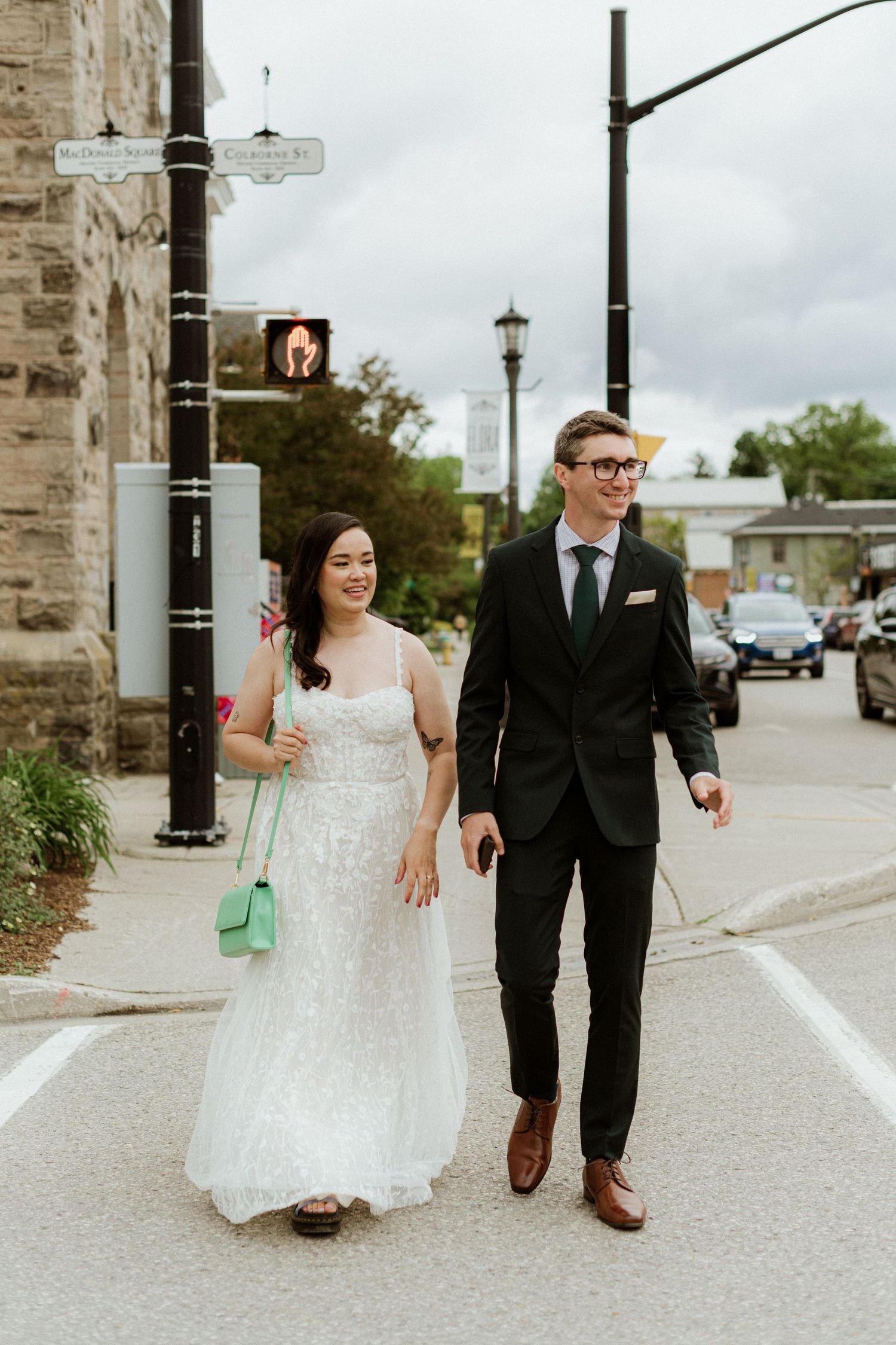 Bride in white lace gown and groom in black suit walking hand in hand across a street intersection.