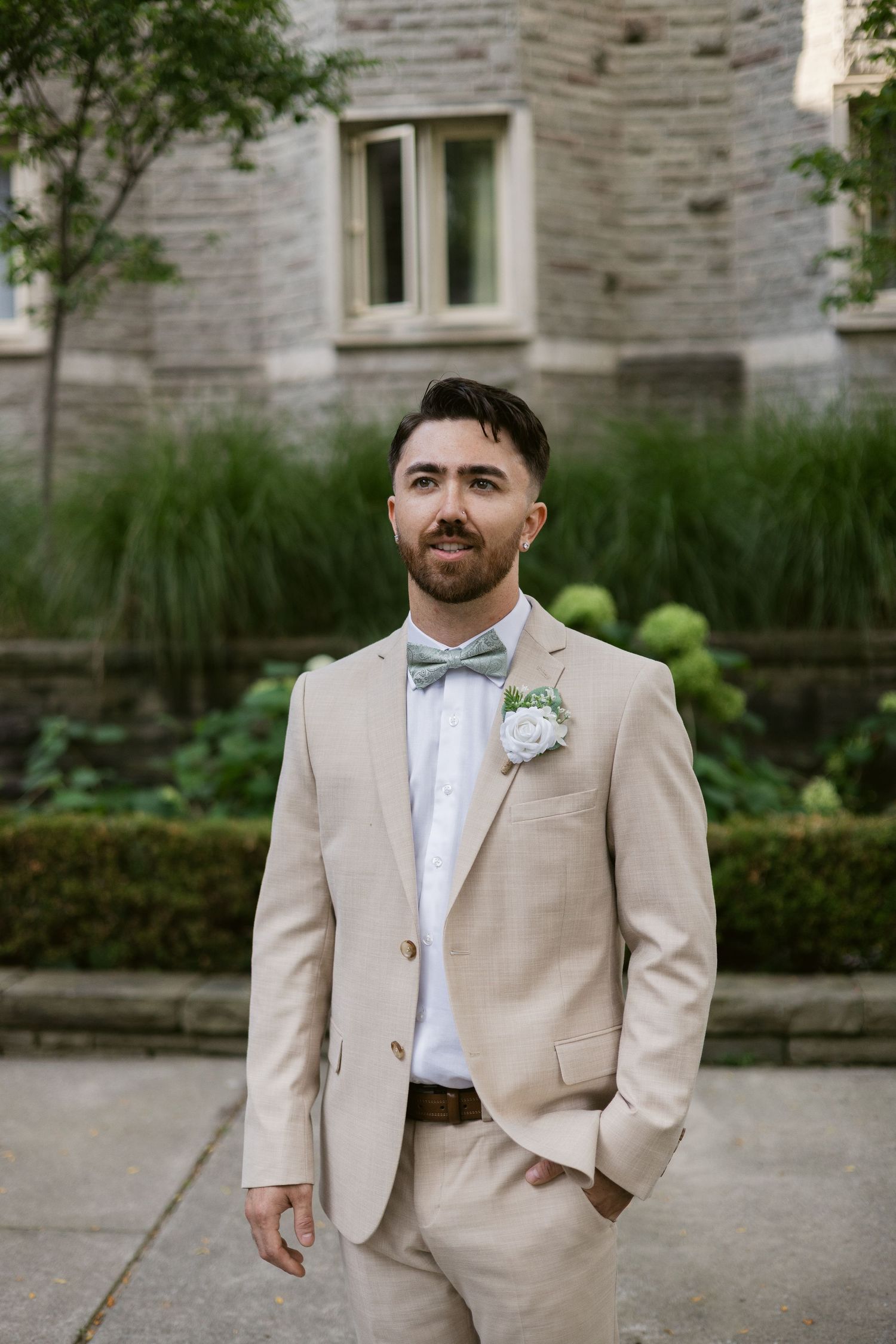 Groom in beige suit with mint bow tie and white boutonniere poses outdoors before stone building.
