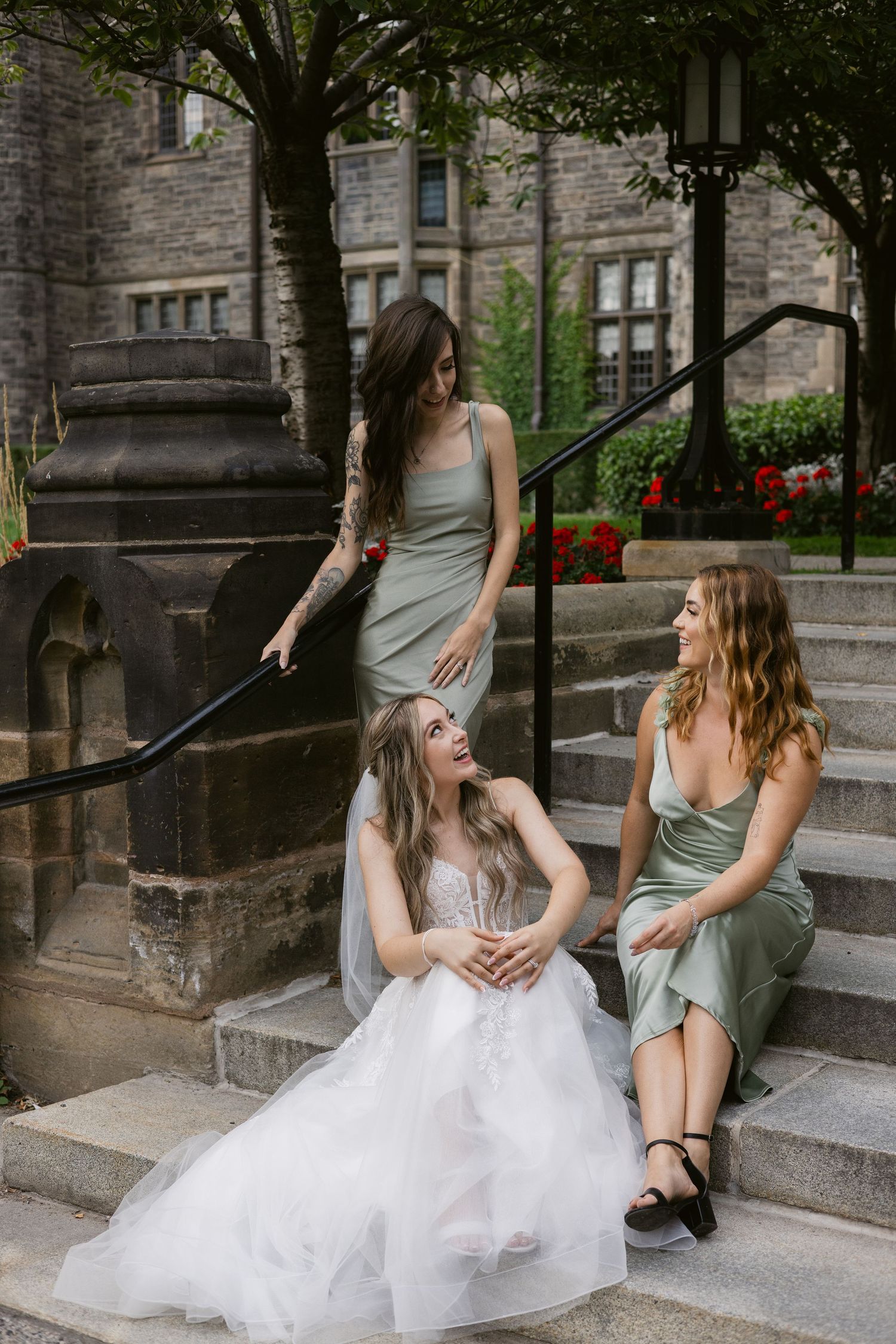 Bride and two bridesmaids in sage green dresses laugh together on stone steps outside historic building.
