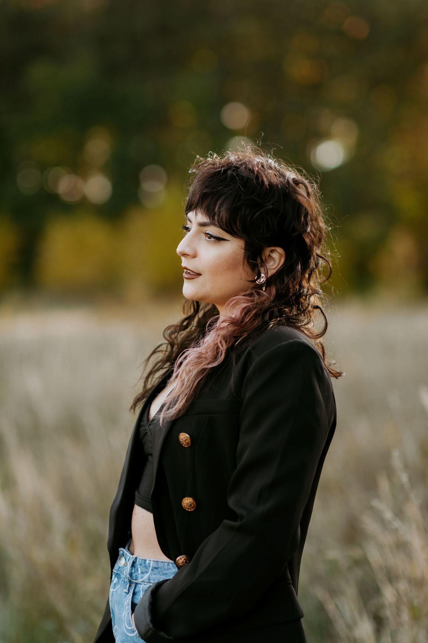 Young woman with curly dark hair and bangs wearing a black blazer and jeans posing in an autumn field.