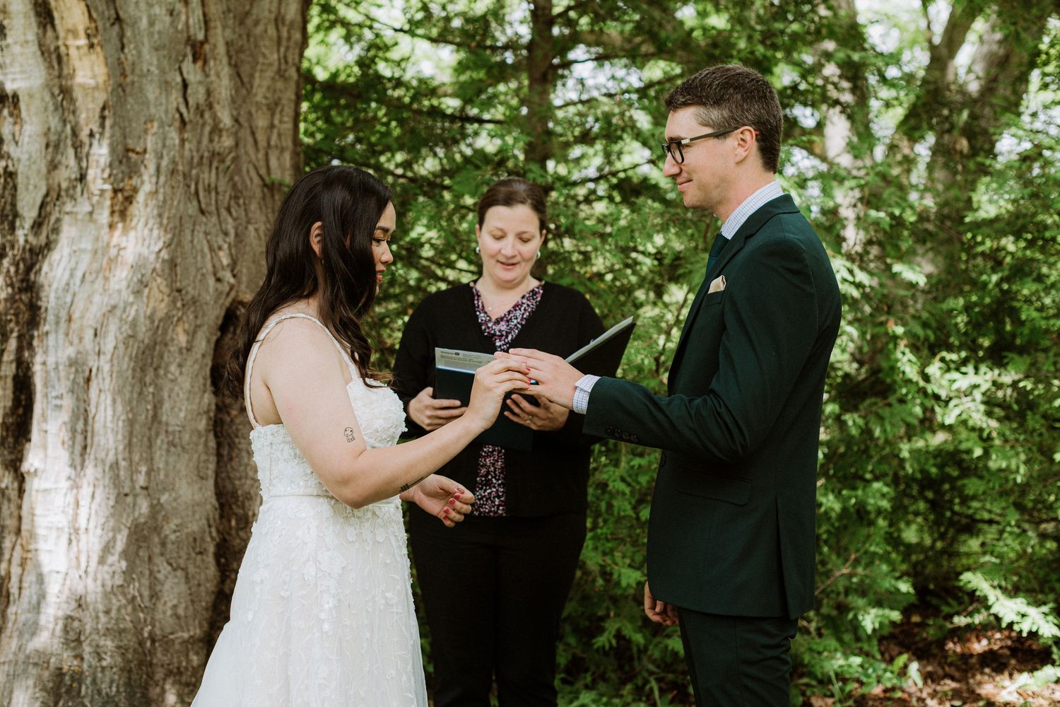 Bride places ring on grooms finger during outdoor wedding ceremony as officiant watches nearby.