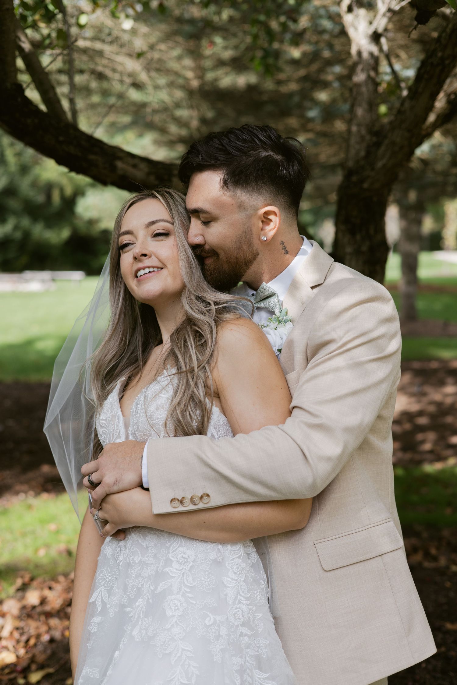 Groom in beige suit embraces smiling bride in lace gown from behind under a shaded garden tree.
