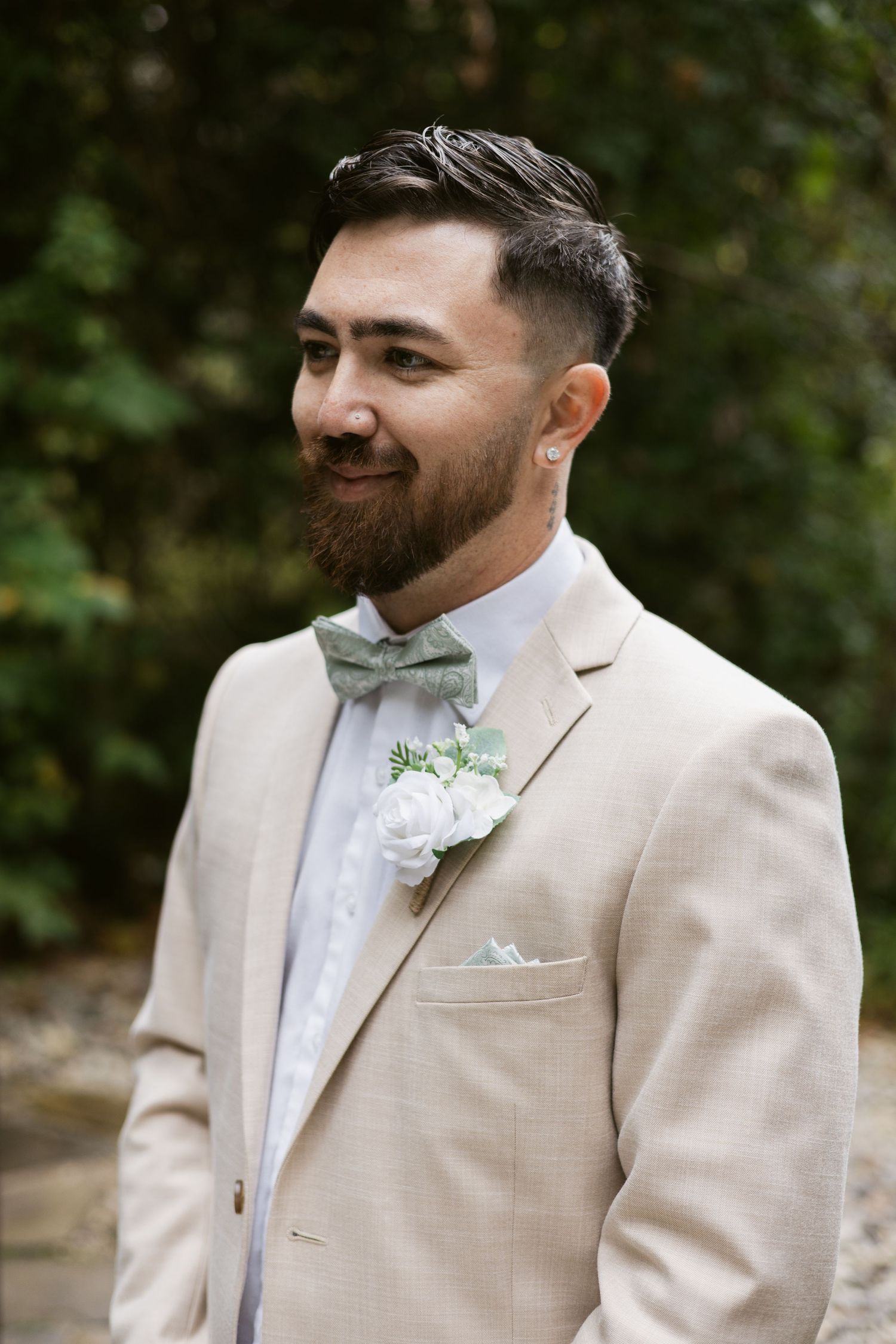 Handsome groom in tan suit with sage green bow tie and white boutonniere smiling outdoors.