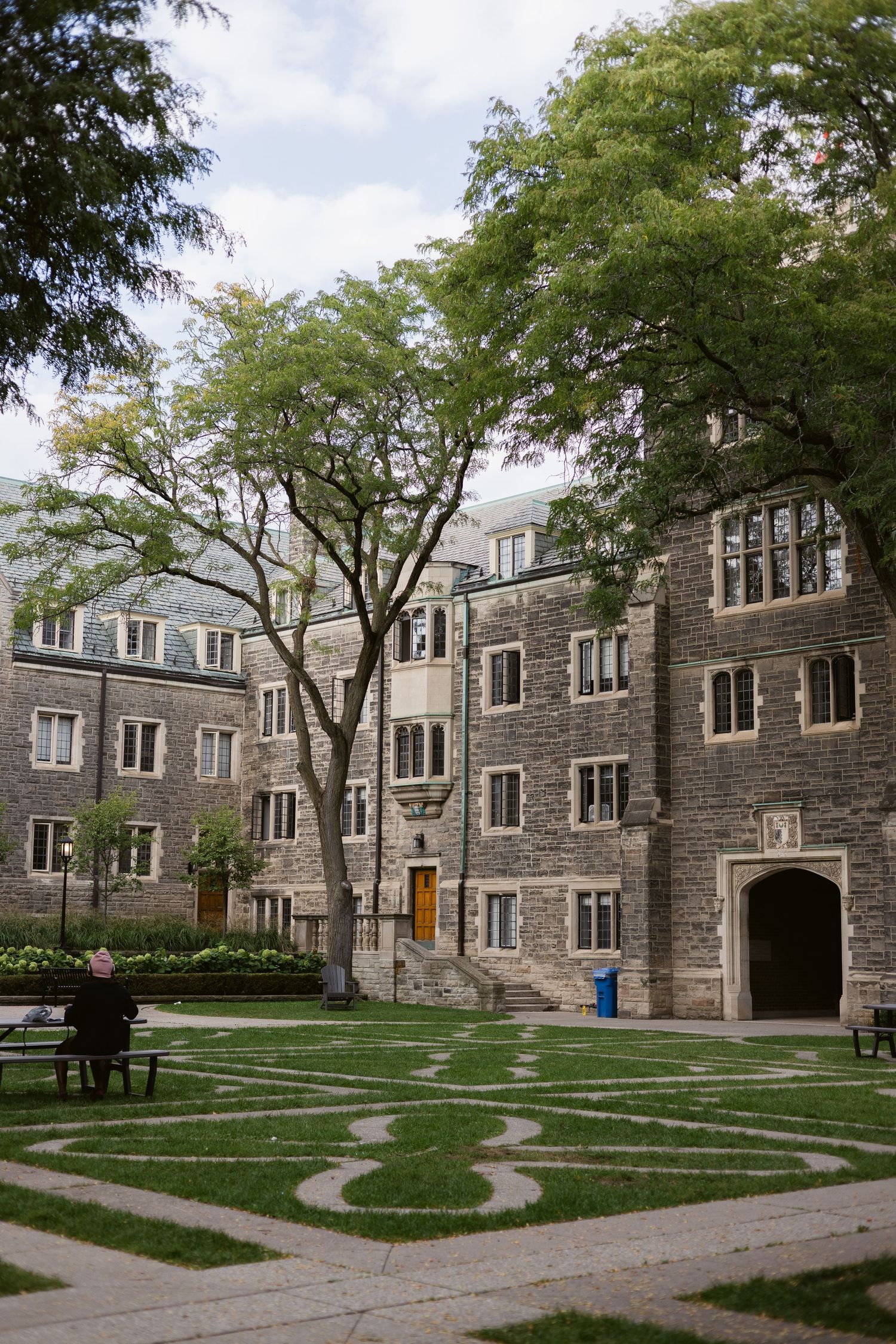 Historic Gothic stone university courtyard with ornate grass labyrinth pattern, mature trees, and a person on a bench.