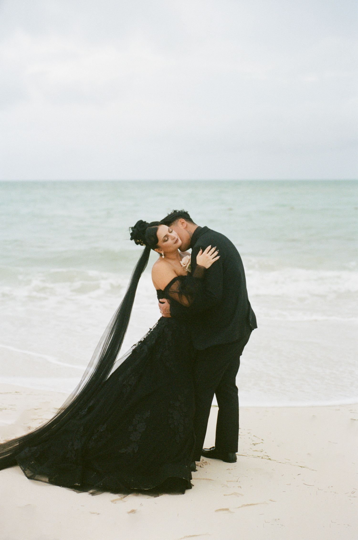 Groom dipping laughing bride in black lace gown with long veil at ocean shoreline, beach wedding portrait.