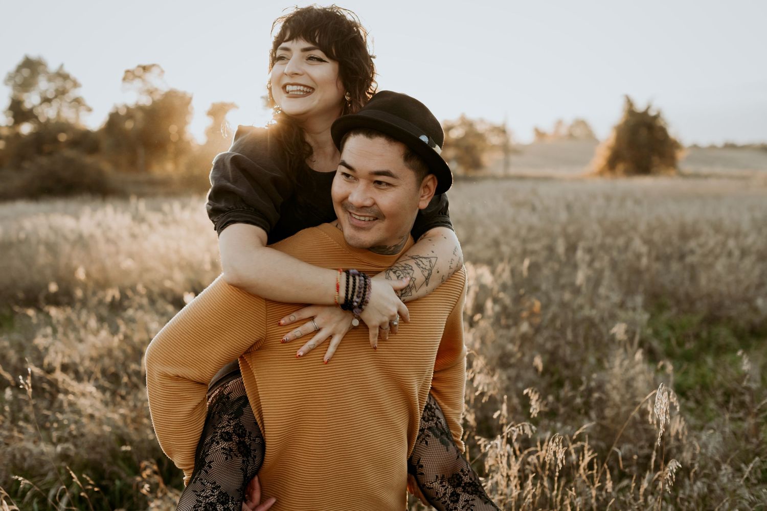 Man in mustard sweater giving laughing woman a piggyback ride through golden sunlit field at sunset.