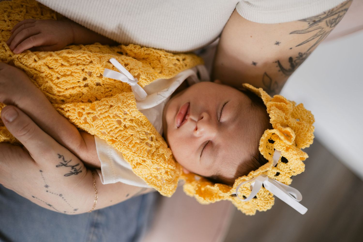 Sleeping newborn baby dressed in yellow crochet outfit and bonnet, cradled in mother's tattooed arms.