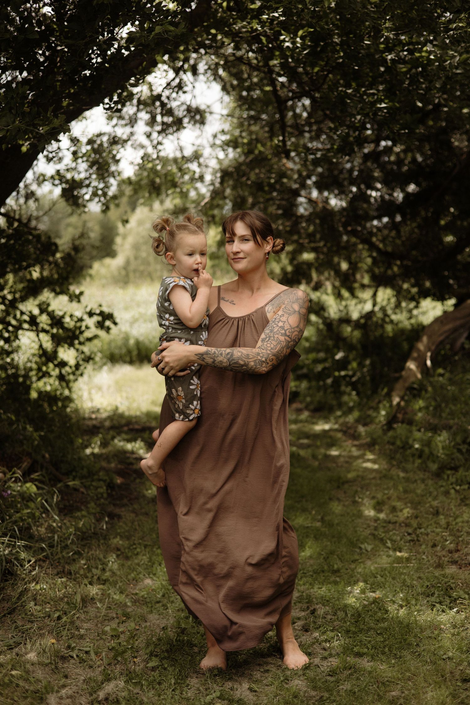 Tattooed mother in brown maxi dress holds toddler girl in daisy romper outdoors in lush green nature setting.