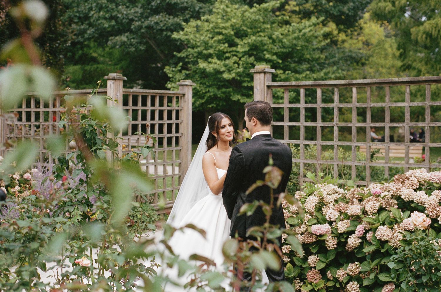 Bride and groom share a romantic moment surrounded by lush garden blooms and wood trellis.