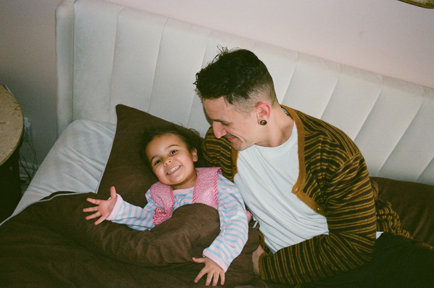 Man in striped cardigan smiles at laughing toddler girl on a white tufted headboard bed.