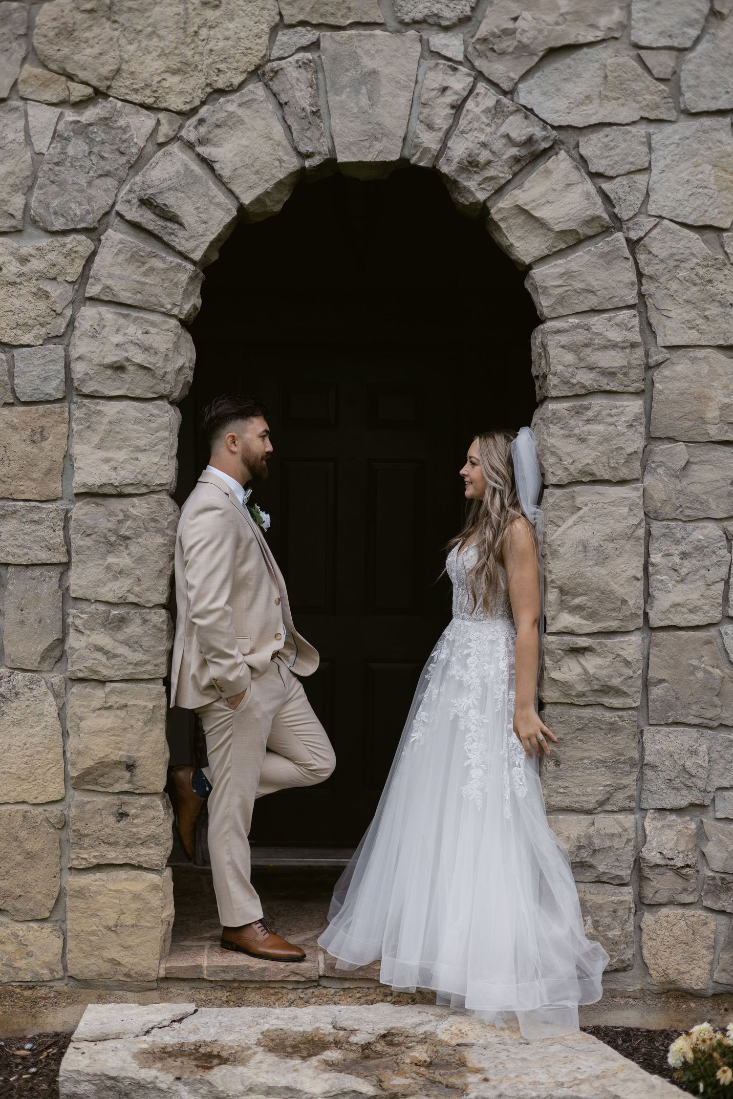 Bride and groom share a romantic moment in a stone archway, groom in beige suit, bride in white lace gown.