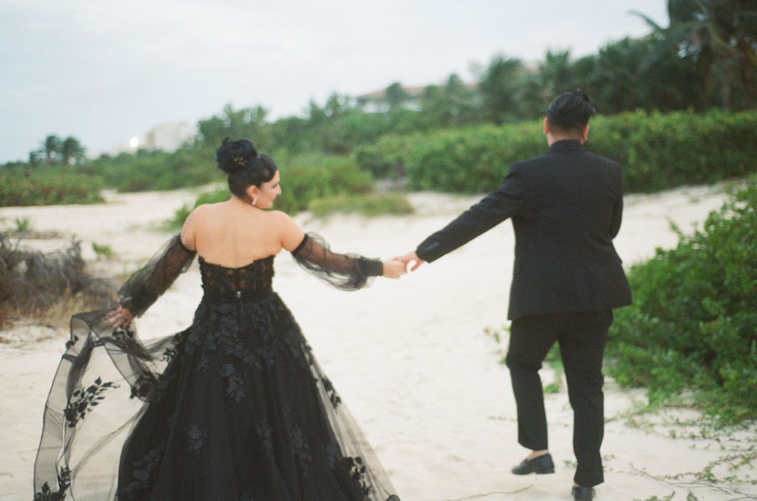 Wedding couple strolls along tropical beach, bride in black lace gown with train flowing in breeze.