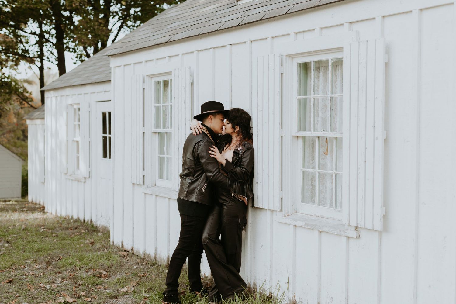 Romantic couple in leather jackets kissing against a white wooden building exterior outdoors.