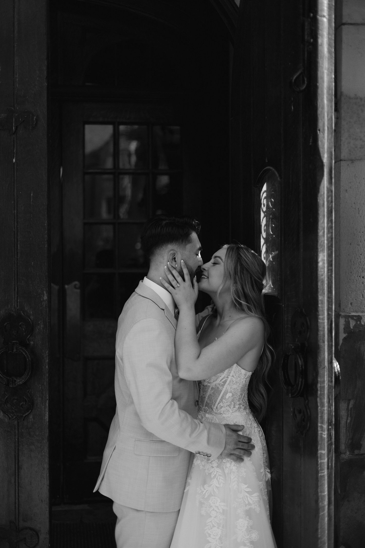 Black and white photo of bride and groom nearly kissing in dramatic wooden doorway of historic venue.