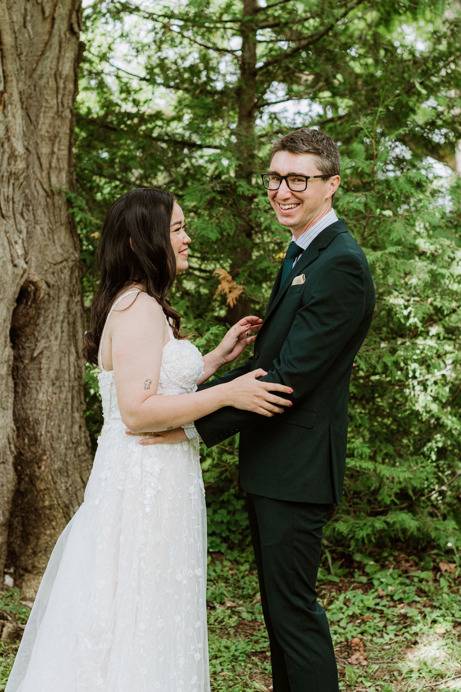 Smiling bride and groom in dark suit laughing together beside large tree in forest setting.