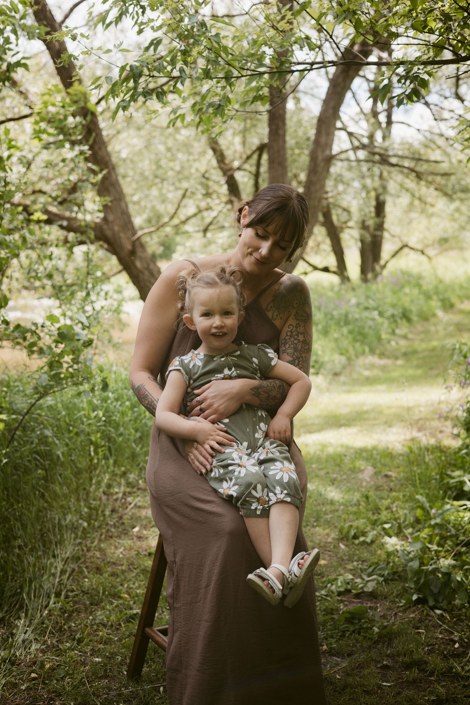 A tattooed mother holds her smiling toddler daughter in a lush green outdoor garden setting.