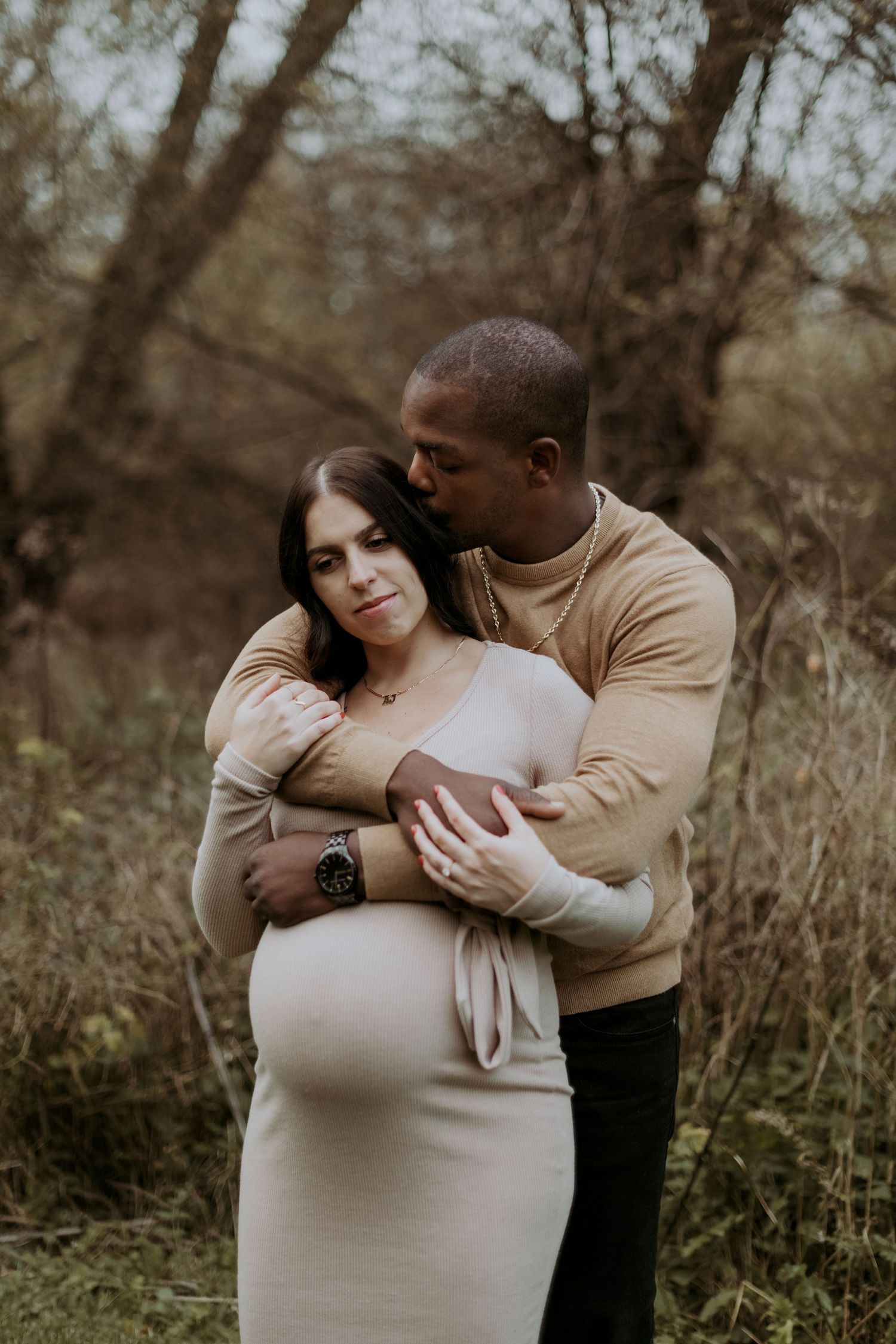 Expectant couple embracing tenderly outdoors, man kissing womans head while holding her baby bump.