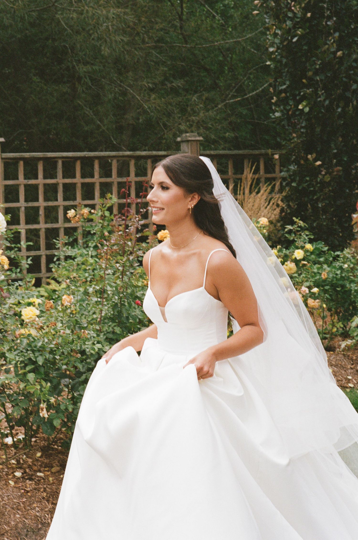 Beautiful bride in a sleeveless white ball gown with veil posing in a garden with yellow roses.