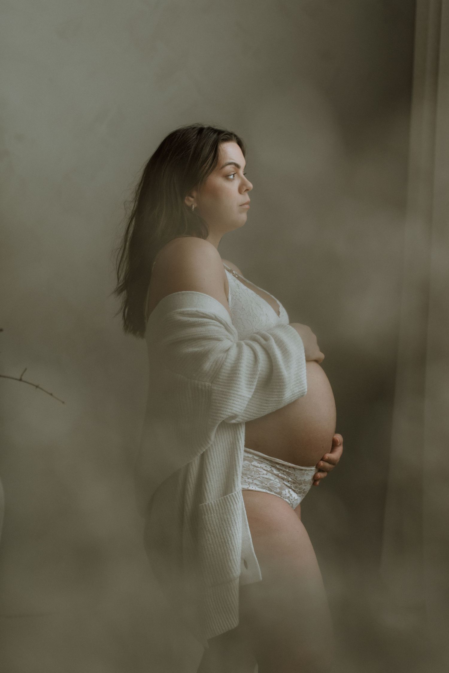 Side profile of a pregnant woman in white lace lingerie and cardigan holding her baby bump.