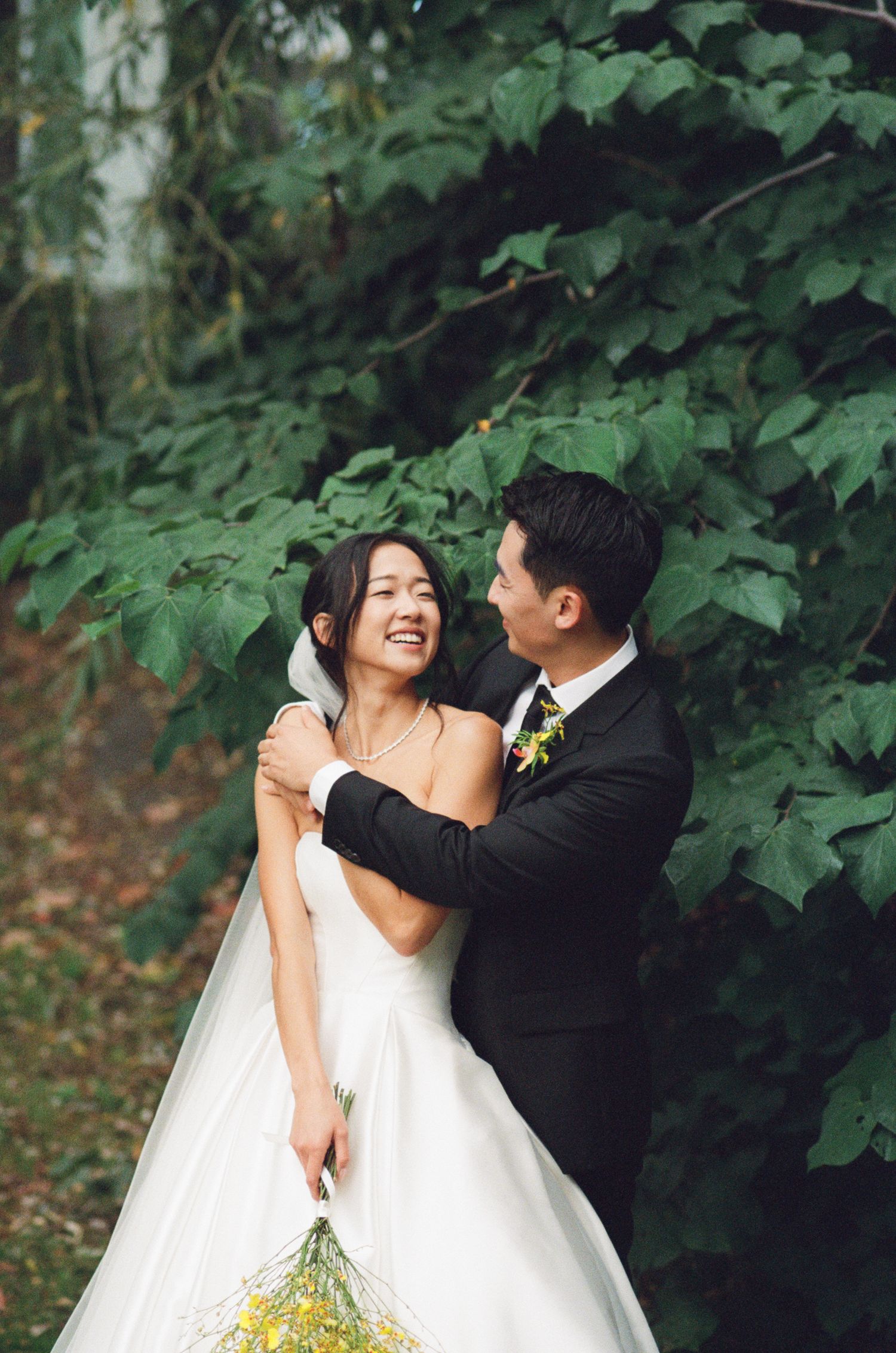 A joyful bride and groom embrace and laugh together outdoors surrounded by lush green foliage.