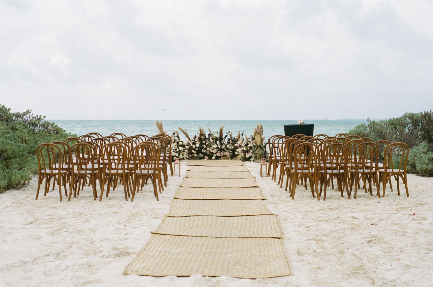 Beach wedding ceremony setup with wooden chairs and woven aisle runner.