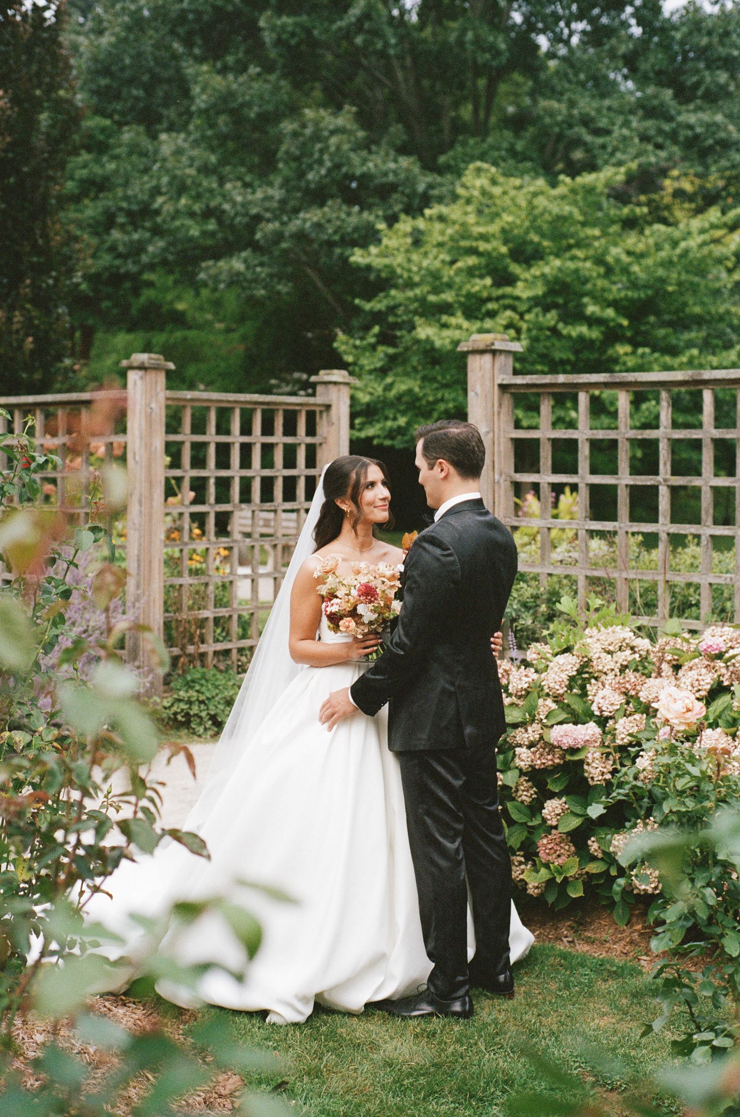 Smiling bride holding bouquet with groom in a romantic garden setting with pink hydrangeas.