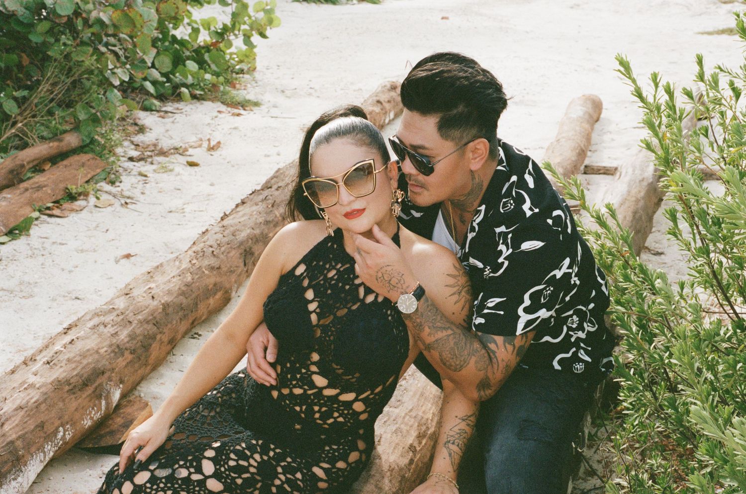Stylish couple in sunglasses sharing intimate moment on sandy beach, man in floral shirt leaning toward woman in black lace.